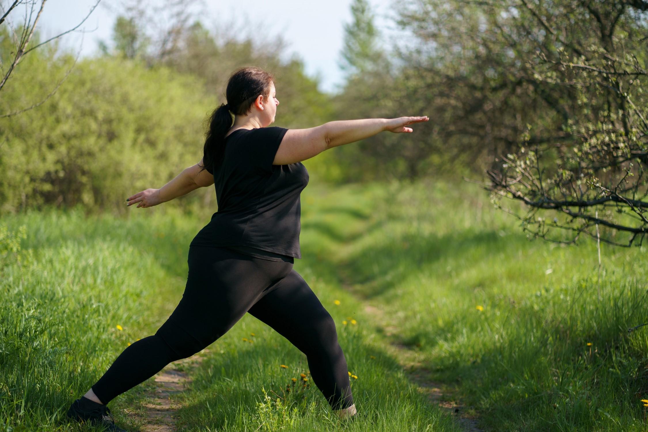 Eine übergewichtige Frau macht die Yoga-Pose der Kriegerin auf einer grünen, mit Bäumen bestandenen Wiese