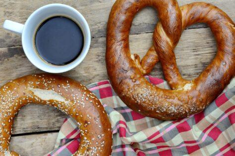 Brezel, Laugenring und eine Tasse schwarzer Kaffee liegen beieinander auf einem Holztisch und zum Teil auf einem karierten Tuch