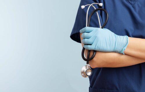 Zu sehen ist der Oberkörper einer Person (zum Teil) mit verschränkten Armen, die einen blauen Arztkittel trägt, hellblaue Handschuhe und ein Stethoskop in der Hand.