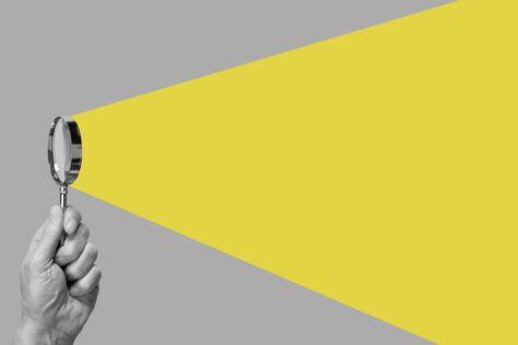 Hand eines Mannes hält eine Lupe, die ein kegelförmiges gelbes Licht ausstrahlt, wie eine Taschenlampe.