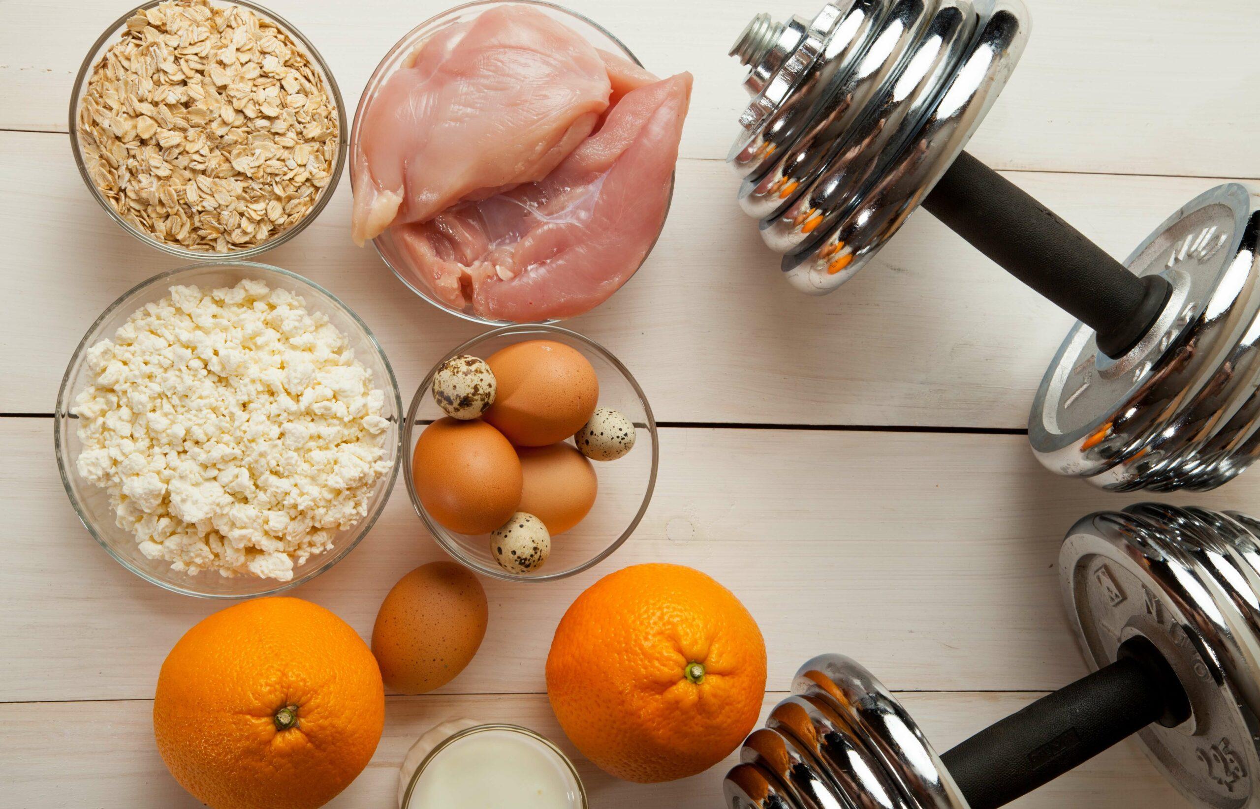 Mehrere Schüsseln sind zu sehen mit Essen (Haferflocken, Eier, Fleisch und Obst) sowie zwei Hanteln.