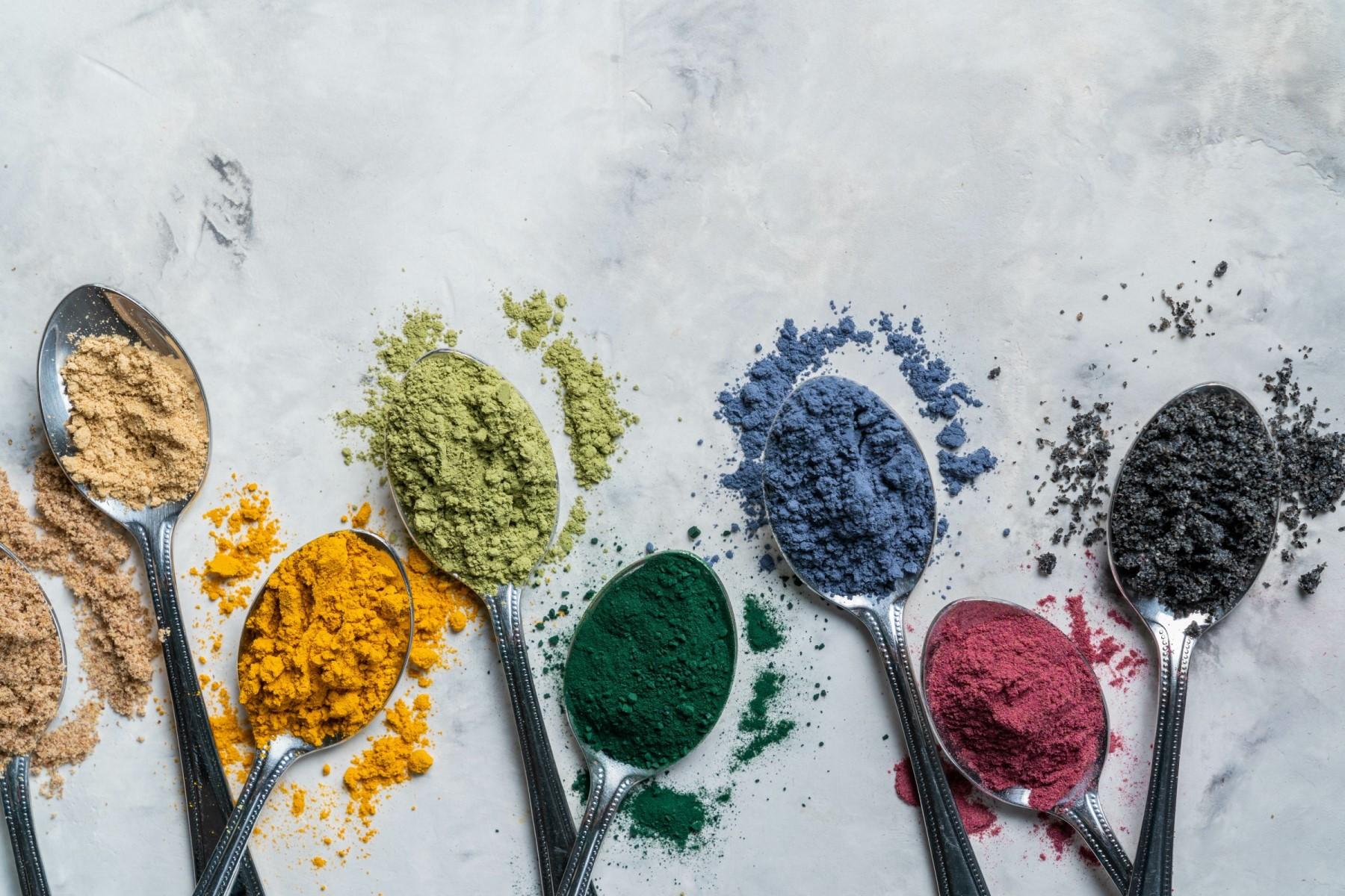 Acht Löffel mit bunten Pulvern darauf liegen auf dem Tisch: Matcha, Kurkuma, Ingwer, Kakao, Spirulina, Chiasamen, Zimt, schwarzer Sesam